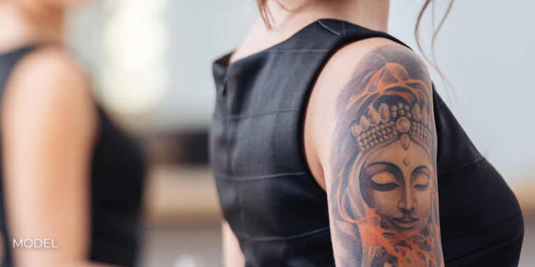 Back view of pretty young woman in black dress with tattoo on her hand standing in front of the mirror