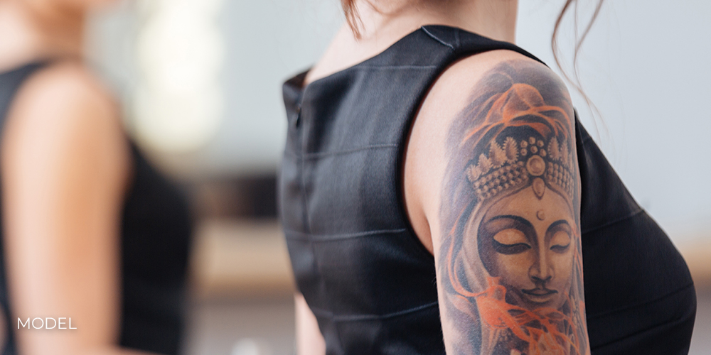Back view of pretty young woman in black dress with tattoo on her hand standing in front of the mirror